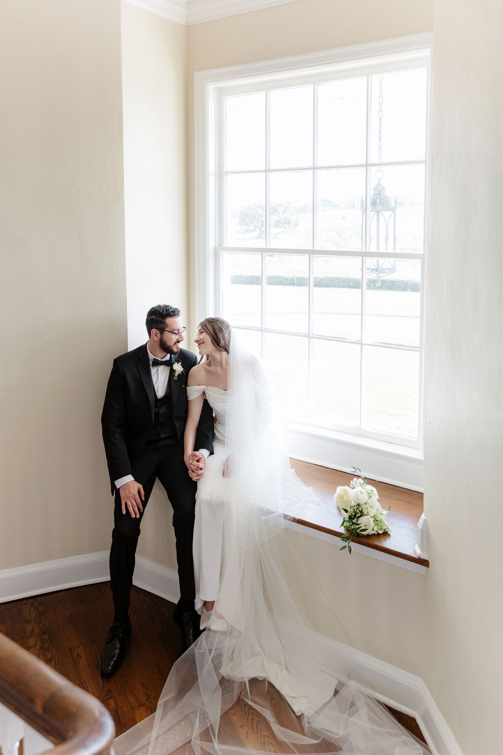 Elegant bride and groom share an intimate moment by a sunlit window, her veil cascading beside a white floral bouquet.