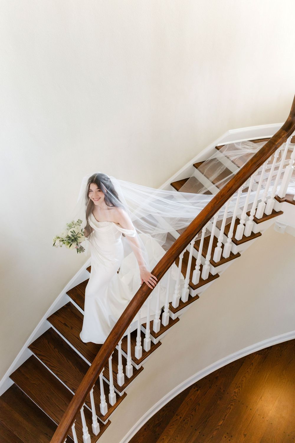 Bride in white off-shoulder gown and flowing veil descends elegant curved staircase holding bouquet.