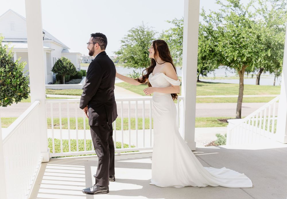 Bride in white gown touches groom's shoulder during first look on white porch at outdoor wedding venue.