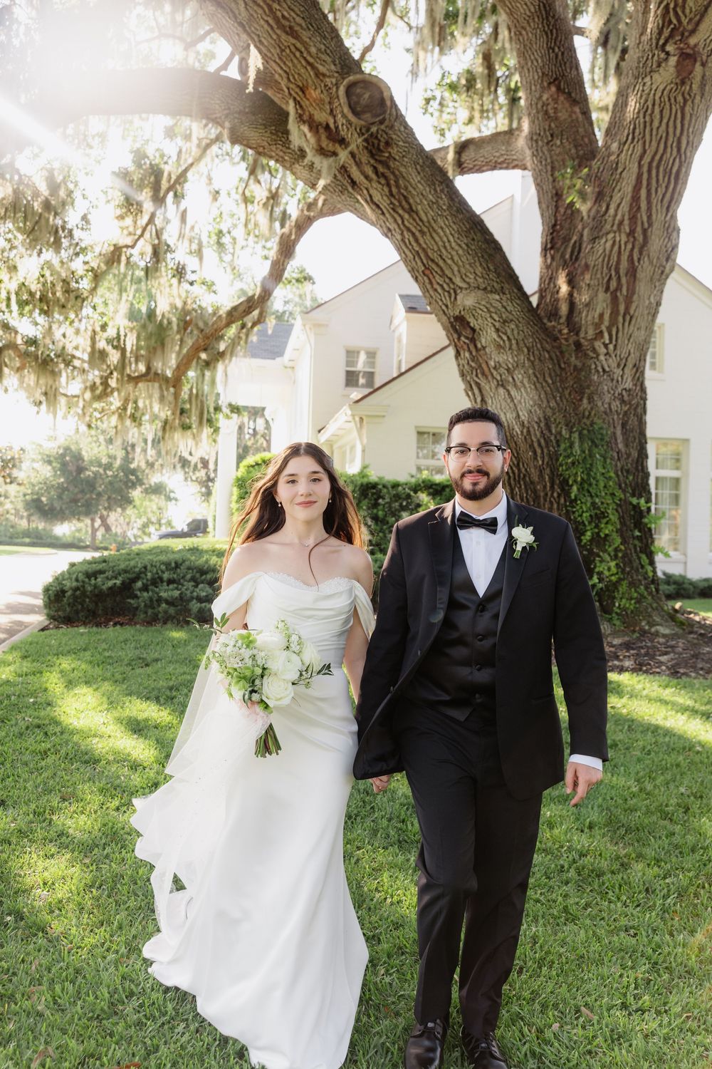 Bride in off-shoulder gown and groom in black tuxedo walk hand-in-hand beneath a Spanish moss oak tree.