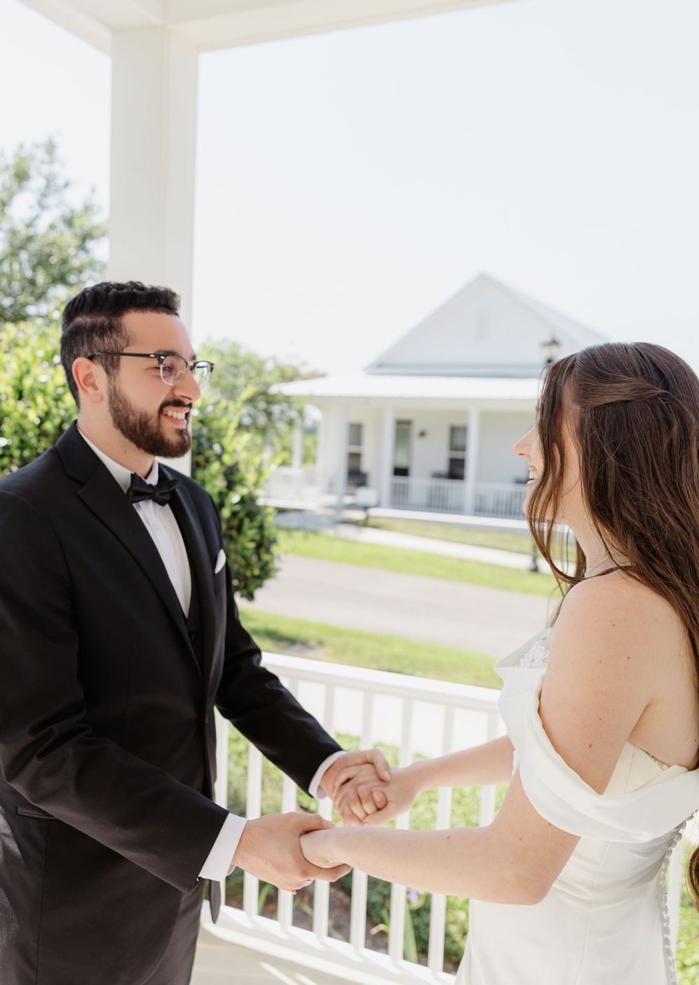 Groom in black tuxedo smiling at bride while holding hands on a white porch during wedding first look.