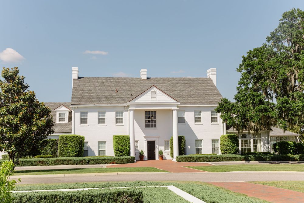 Elegant white colonial mansion with manicured hedges and brick pathway on a sunny day.