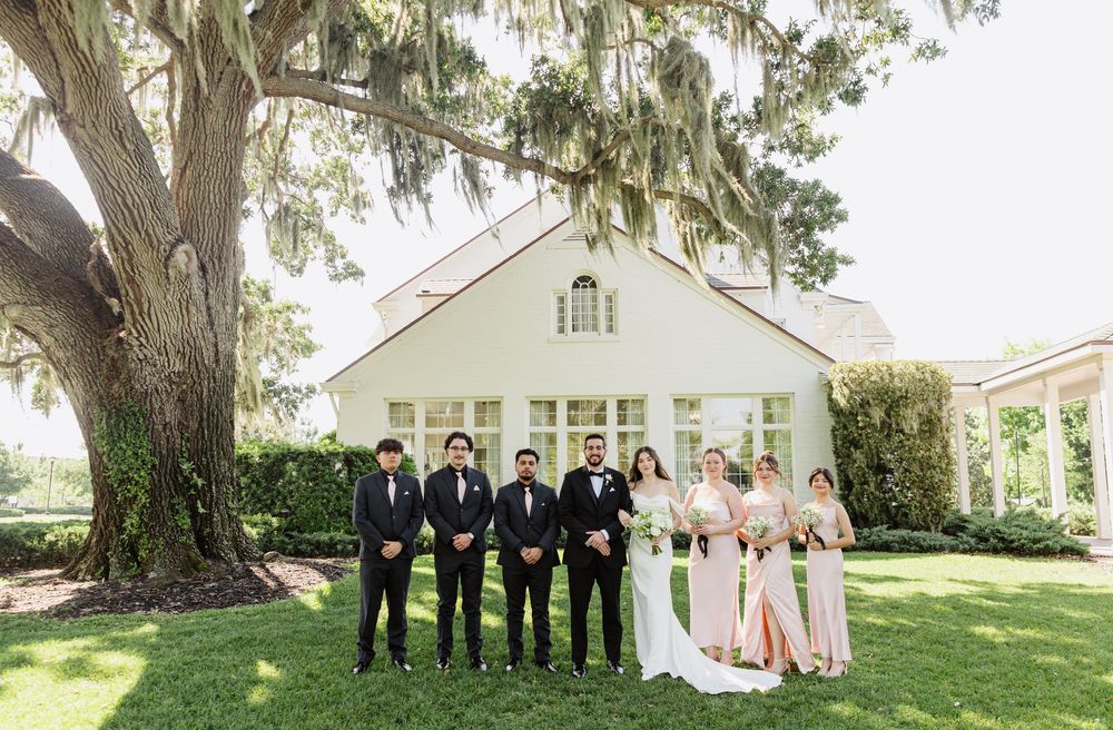 Wedding party posing in formal attire on green lawn beside a large oak tree and white chapel.