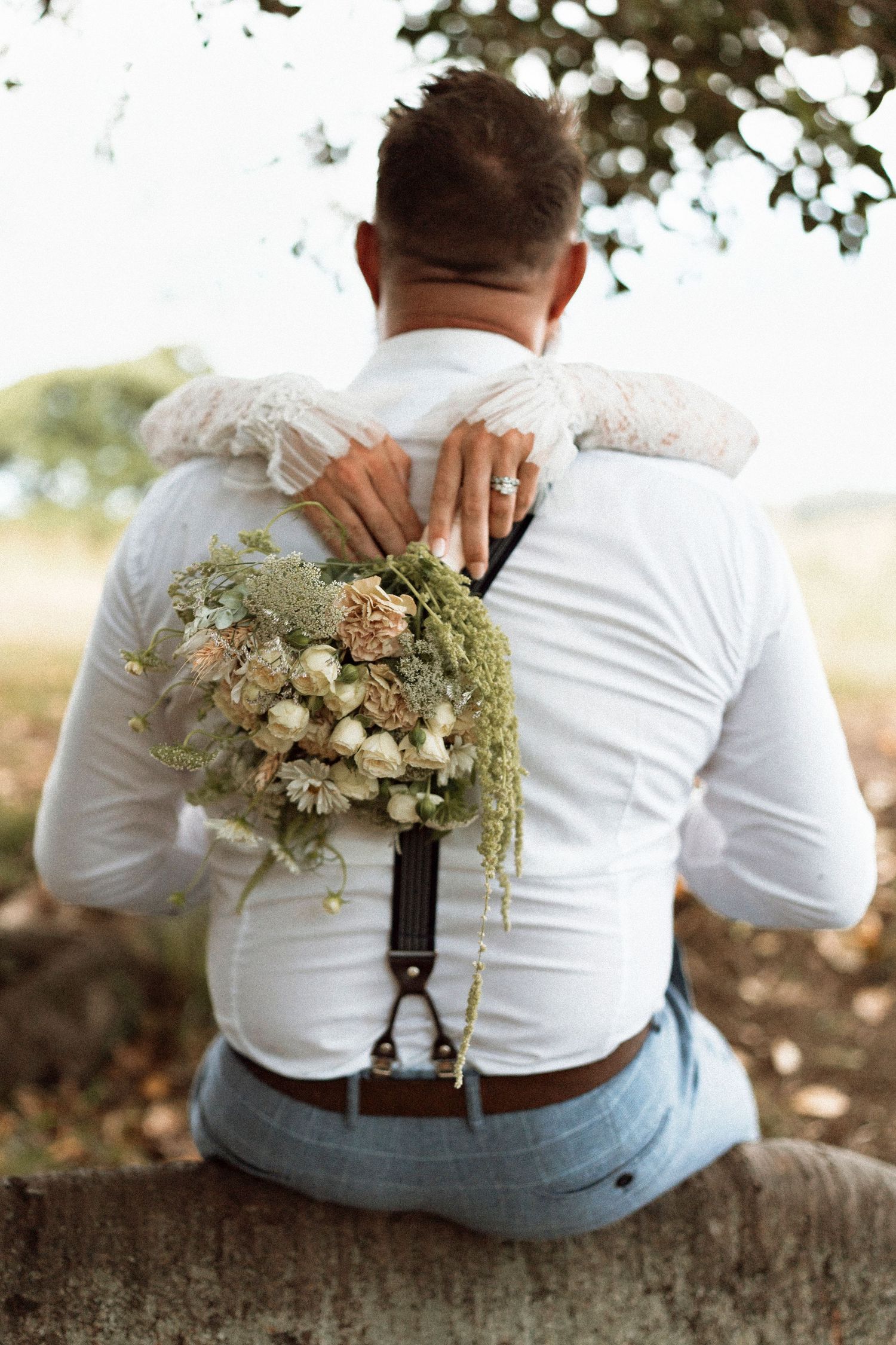 Bride embracing groom from behind, holding a rustic bouquet, both in wedding attire, seated outdoors.