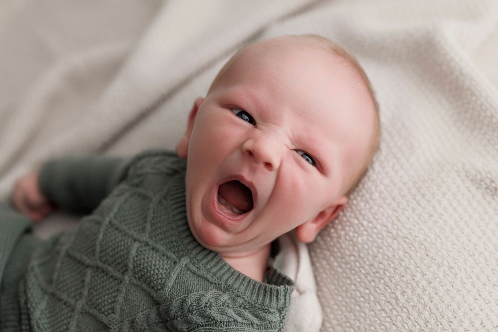 Adorable baby in green knit sweater yawning while lying on white blanket, newborn photography portrait.
