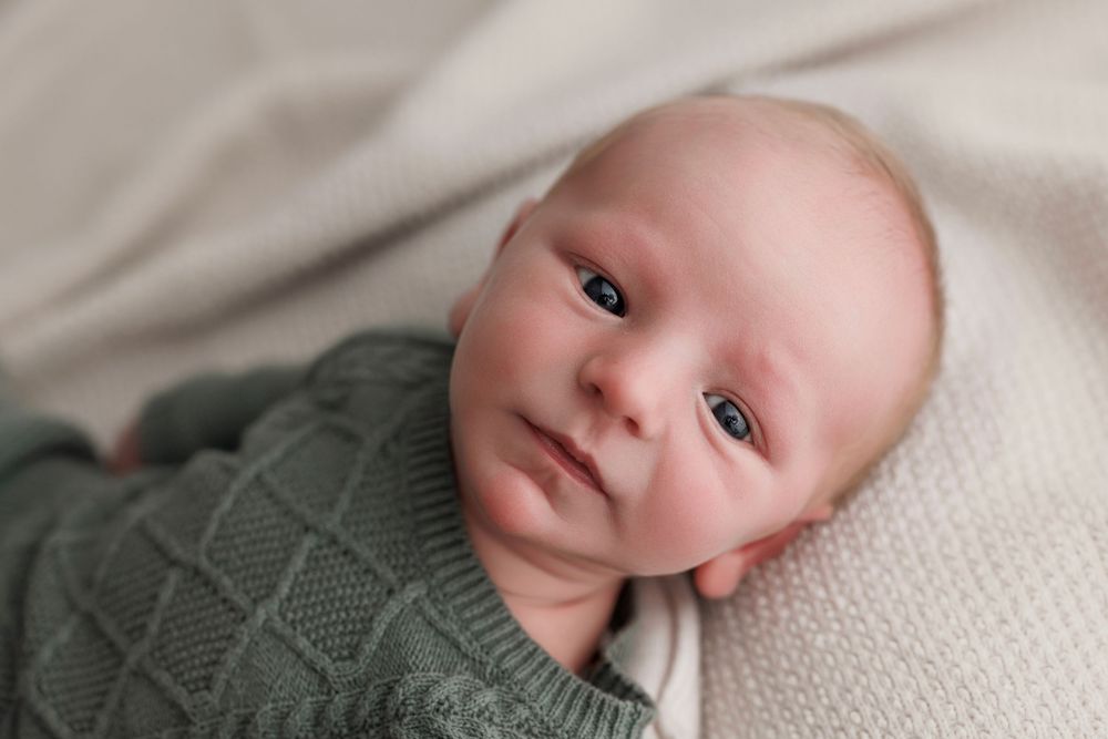 Newborn baby in green knit sweater lying on white blanket, looking up with bright blue eyes.