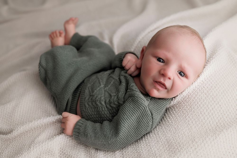 Awake newborn baby in green knit romper lying on white blanket, looking at camera.