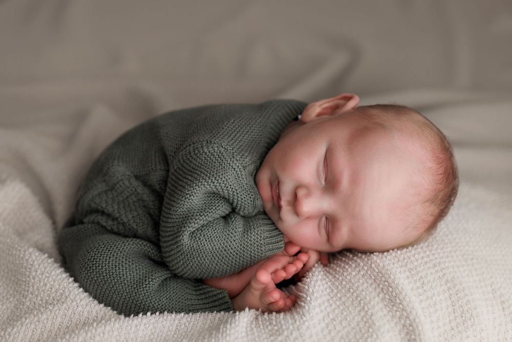Newborn baby sleeping peacefully in a green knit romper on a soft white blanket.
