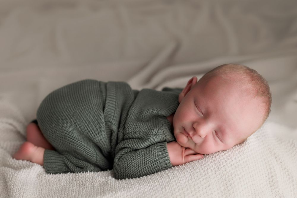 Newborn baby sleeping peacefully in green knit romper on white blanket.