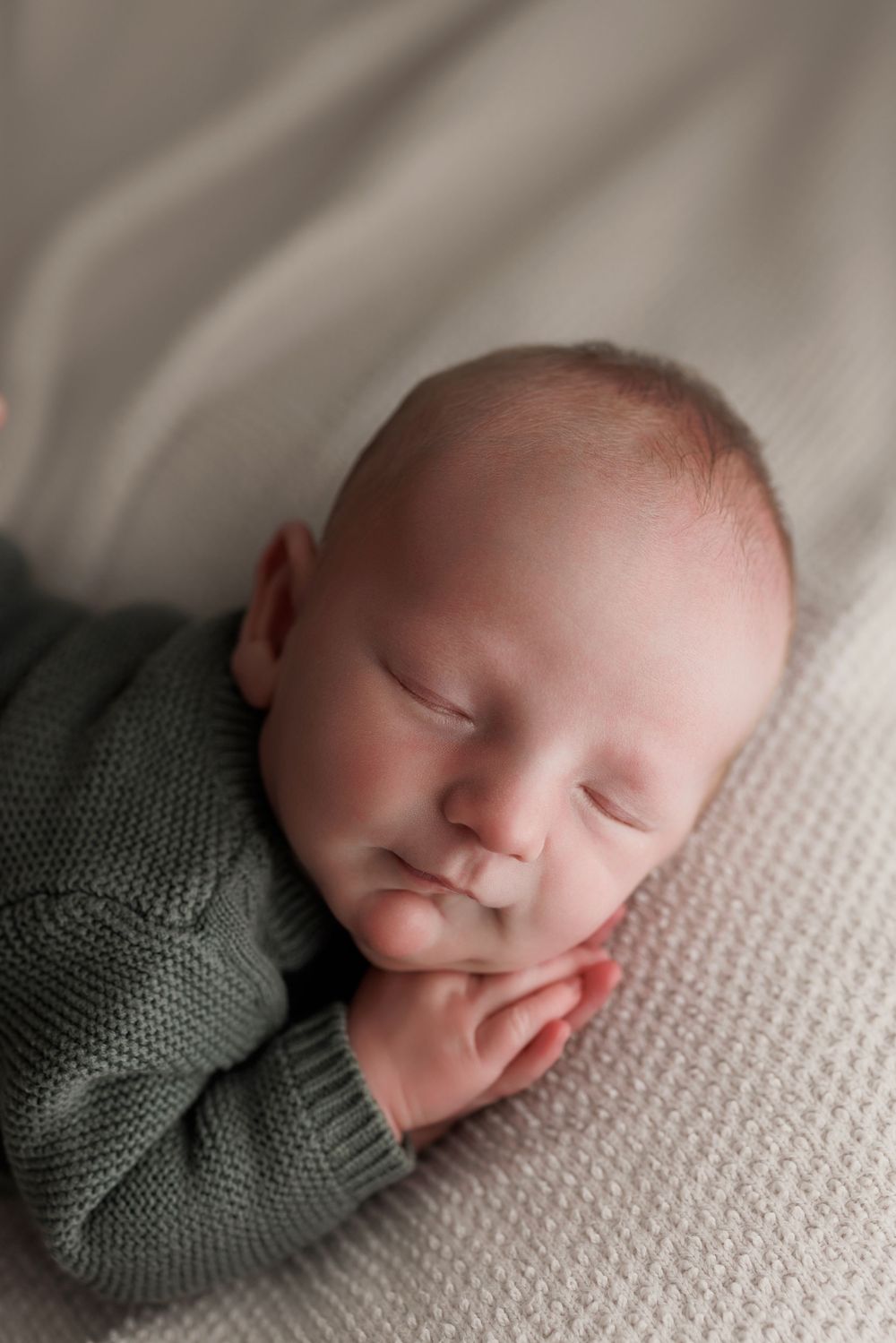 Sleeping newborn baby in green knit sweater resting on white textured blanket.