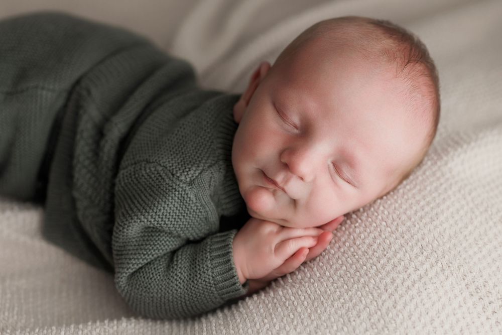 Sleeping newborn baby in green knit romper curled up peacefully on soft white blanket.