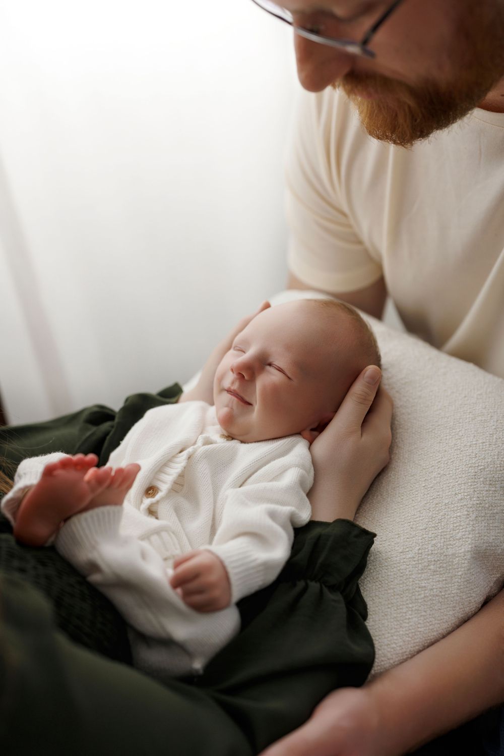 A bearded father in glasses tenderly holds his sleeping newborn baby dressed in white.