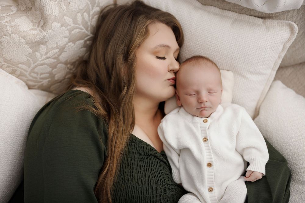 A mother gently kisses her sleeping newborn baby dressed in white, both resting on soft pillows.