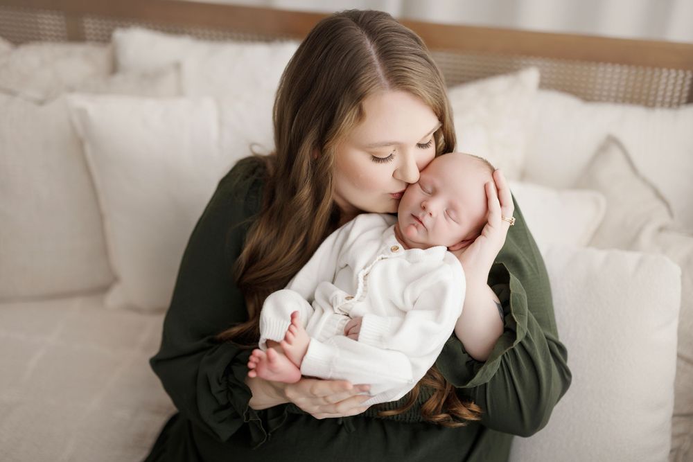 A mother in a green dress tenderly kisses her sleeping newborn baby wrapped in white.
