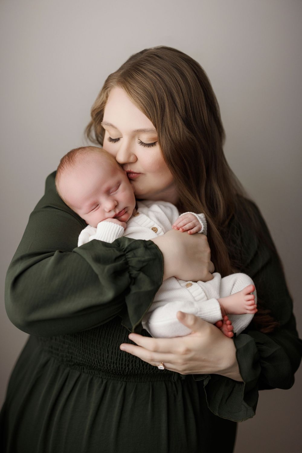 Mother in dark green dress tenderly kissing her sleeping newborn baby.