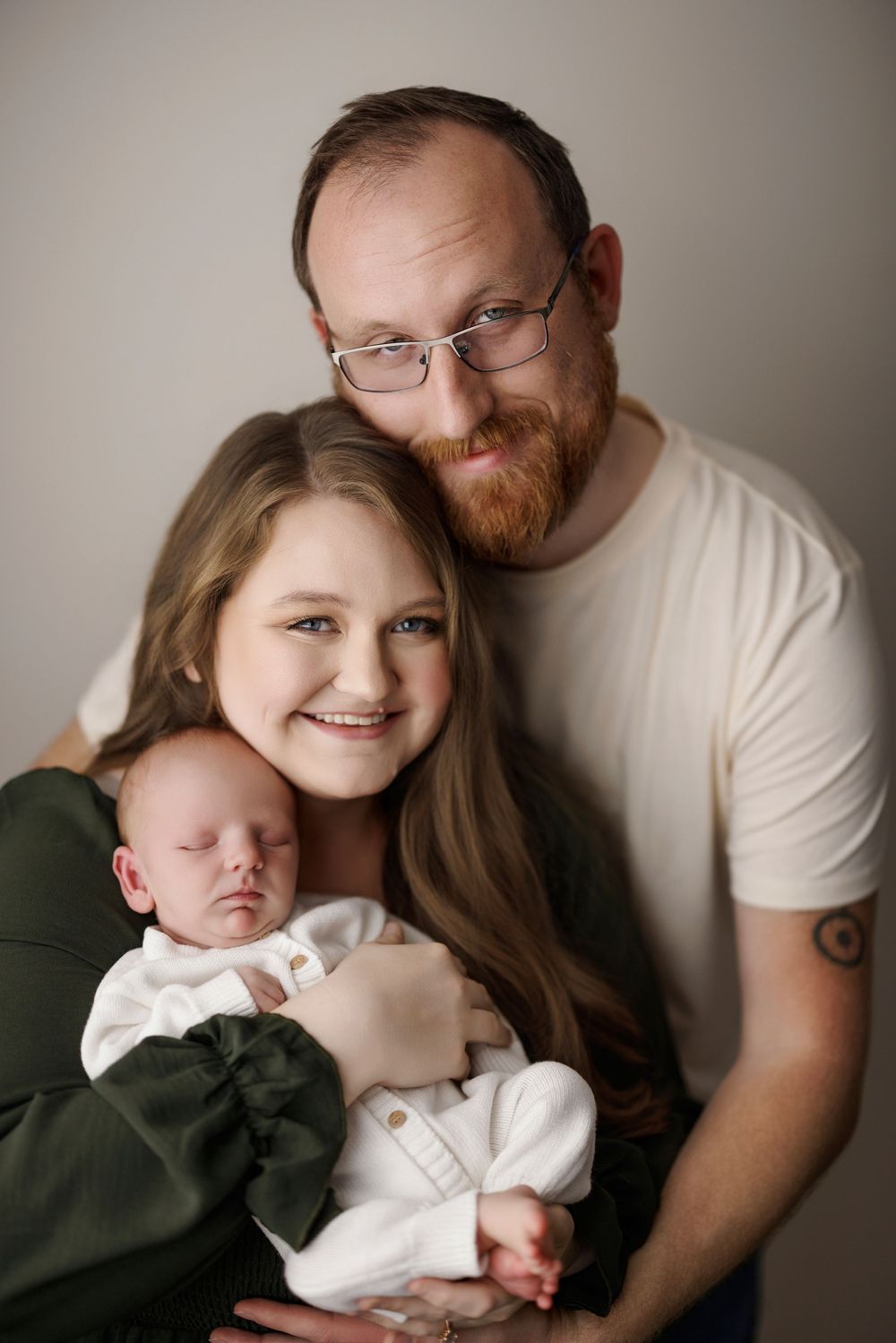 Smiling young mother in green dress holds sleeping newborn baby while bearded father with glasses leans in close.