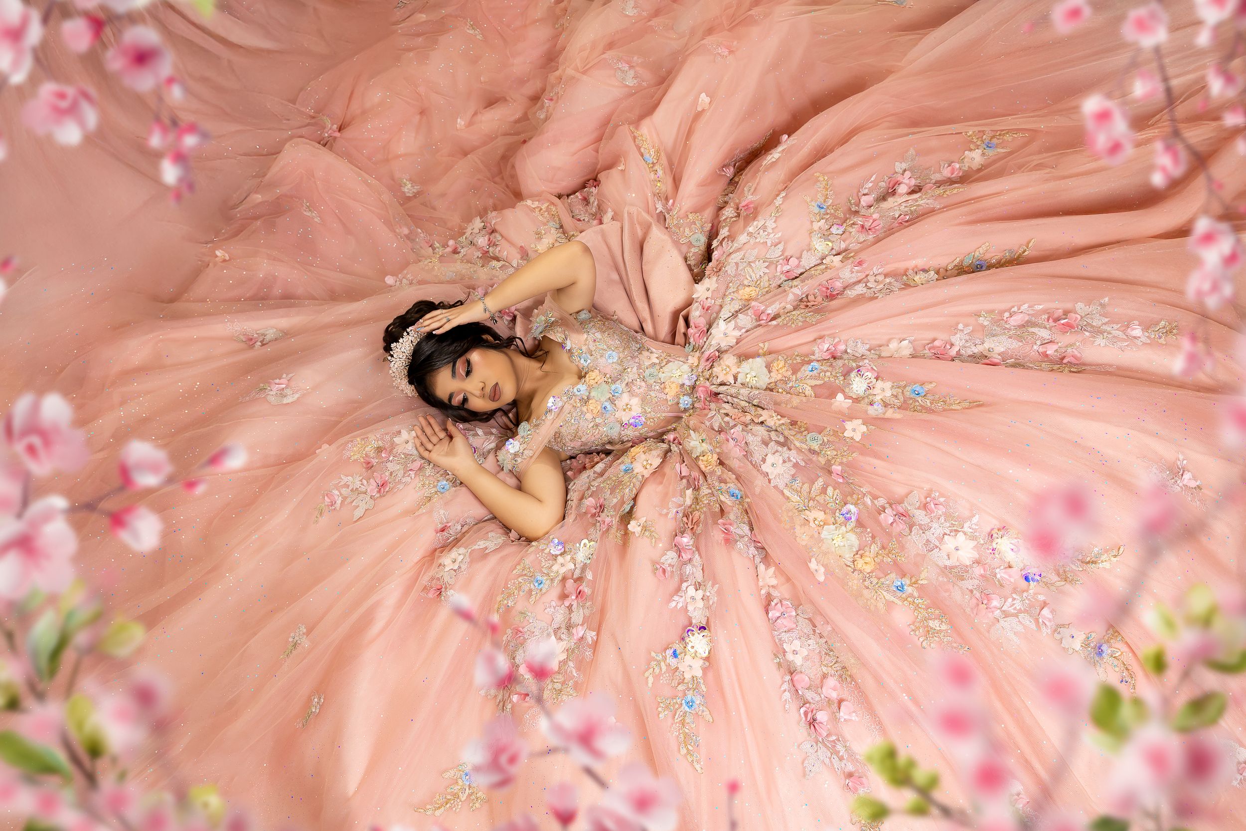 Young woman in a blush pink floral ballgown lying gracefully among cherry blossoms in an ethereal portrait.