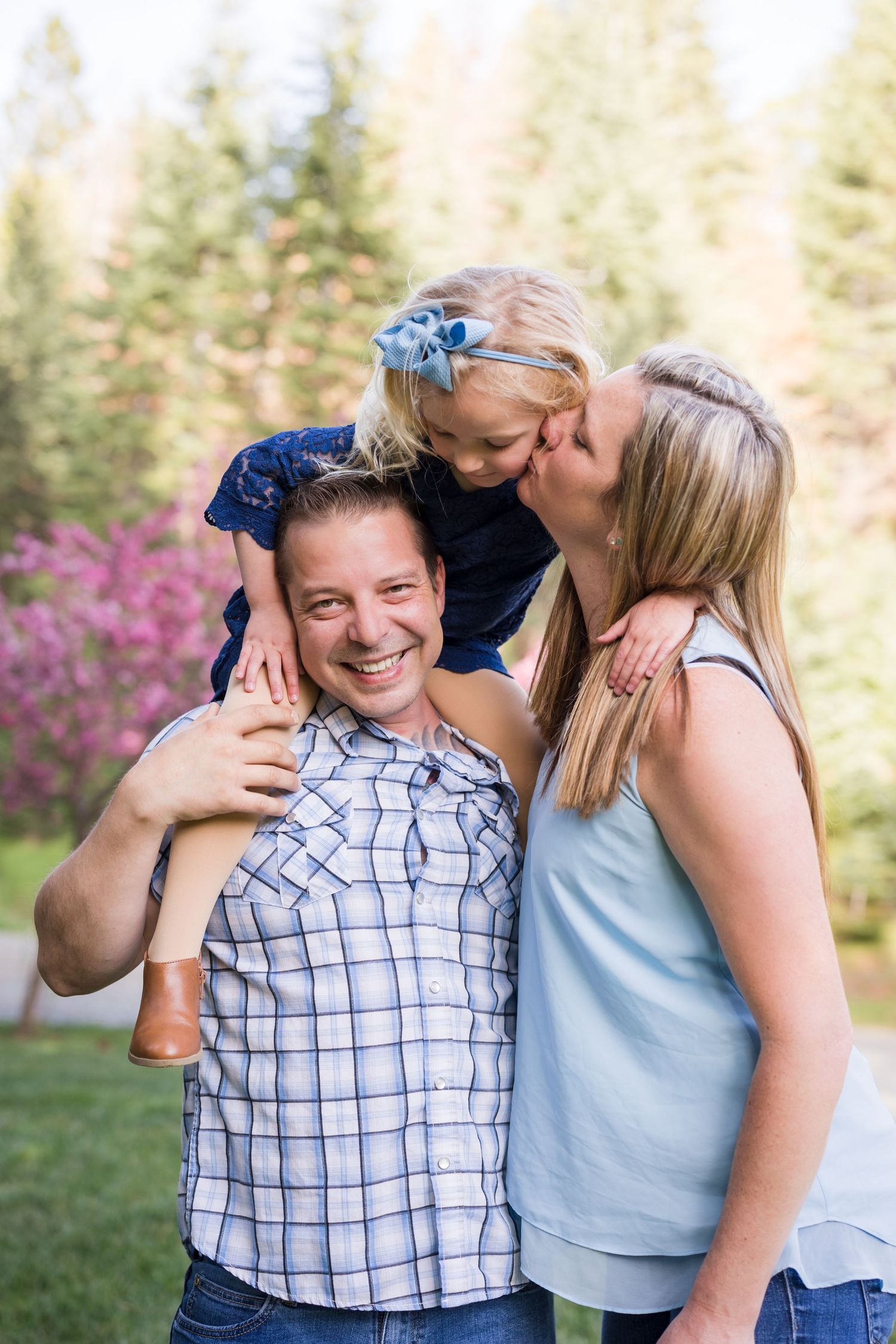 Happy family moment with dad carrying daughter on shoulders while mom kisses her cheek outdoors.