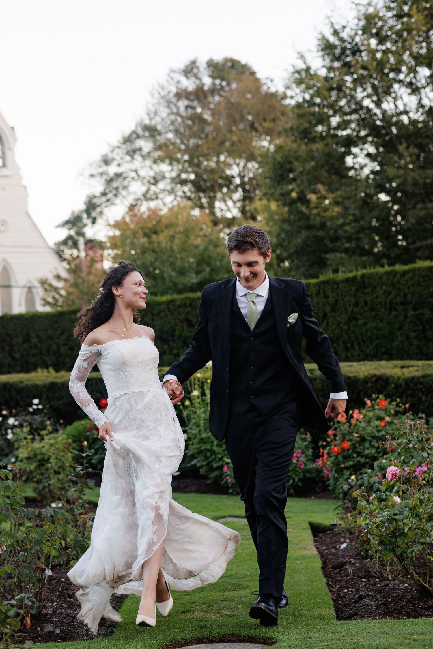 Bride in white lace gown and groom in black suit walk hand-in-hand through a lush garden on their wedding day.