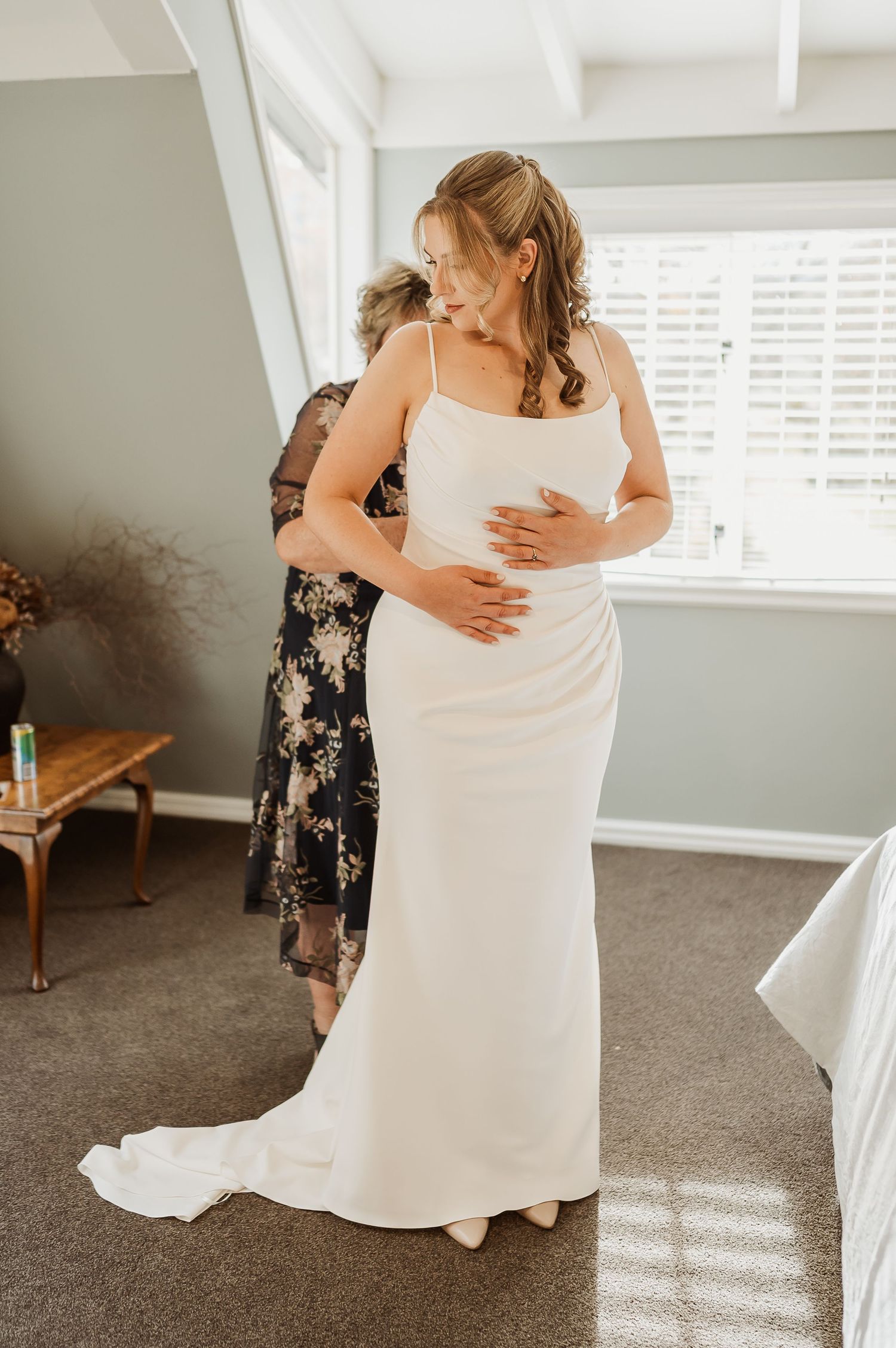 Bride in elegant white spaghetti strap wedding gown being helped to dress in a bright room.