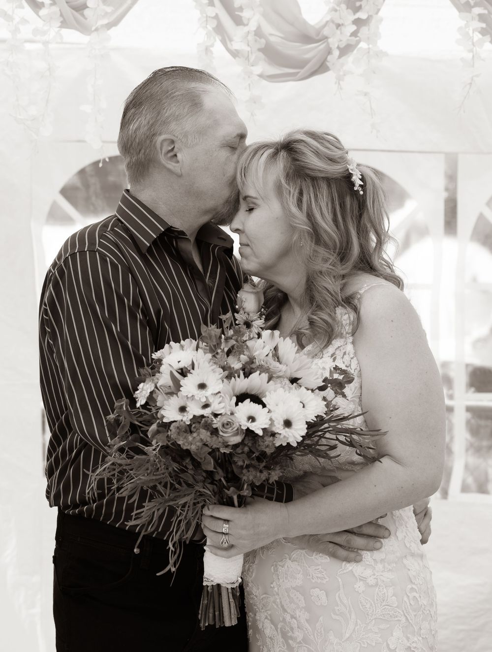 Black and white photo of a groom kissing bride holding a bouquet of daisies at a wedding ceremony.