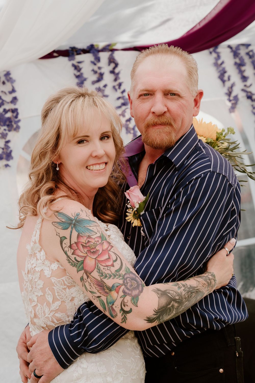 A smiling couple with tattoos embraces at their wedding, the bride in lace and groom in a navy striped shirt, under floral decor.