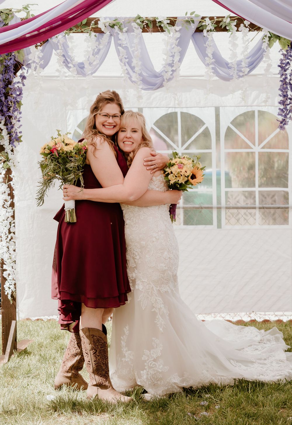 Bride in white lace gown hugs bridesmaid in burgundy dress holding bouquets under floral wedding arch.