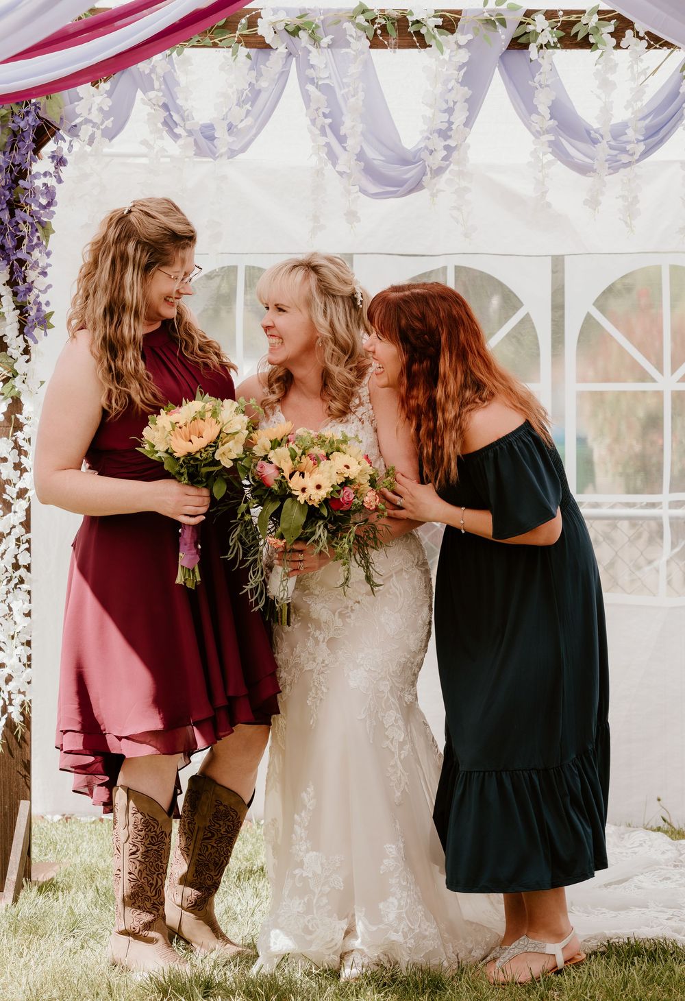 Bride in white lace gown laughs with two women in burgundy and teal dresses, holding floral bouquets at outdoor wedding.