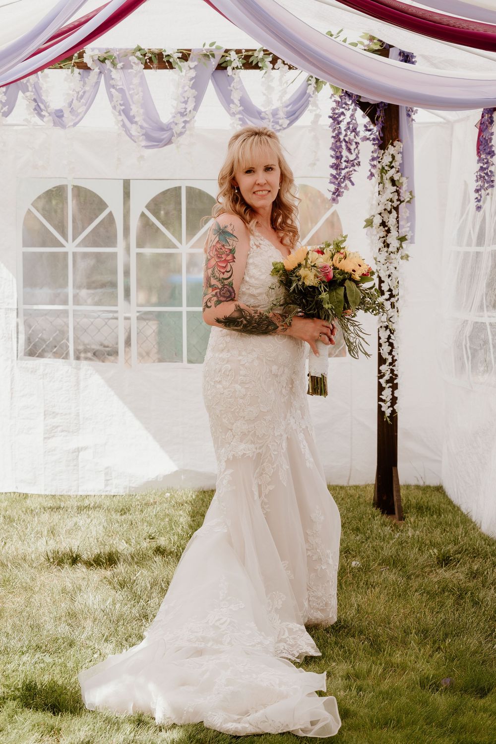 Bride in lace mermaid wedding gown holding floral bouquet stands under decorated canopy with purple draping outdoors.