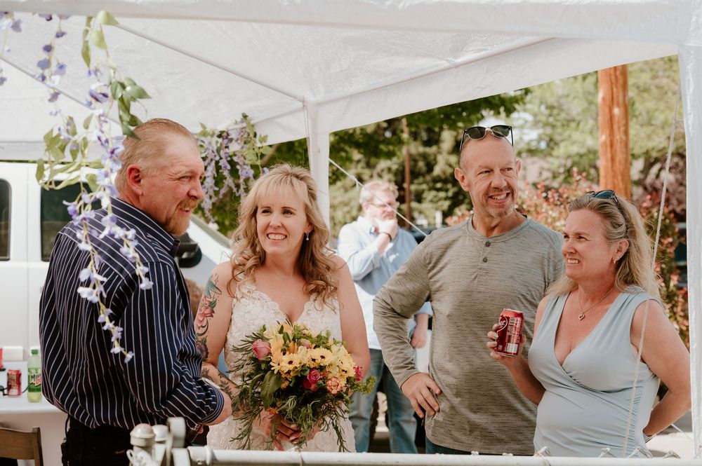 Bride holding bouquet laughs with family under white canopy at outdoor wedding reception.