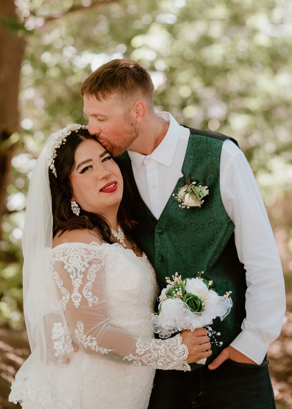 Bride in lace gown holding white bouquet as groom in green vest kisses her cheek outdoors.