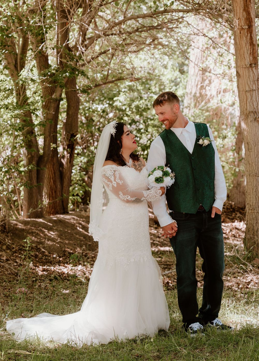 Bride in white lace gown and groom in green vest holding hands and smiling at each other outdoors among trees.