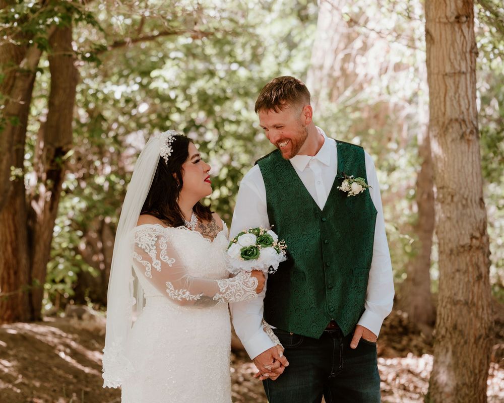 Bride and groom holding hands and smiling at each other outdoors, wearing white lace gown and green vest with boutonniere.