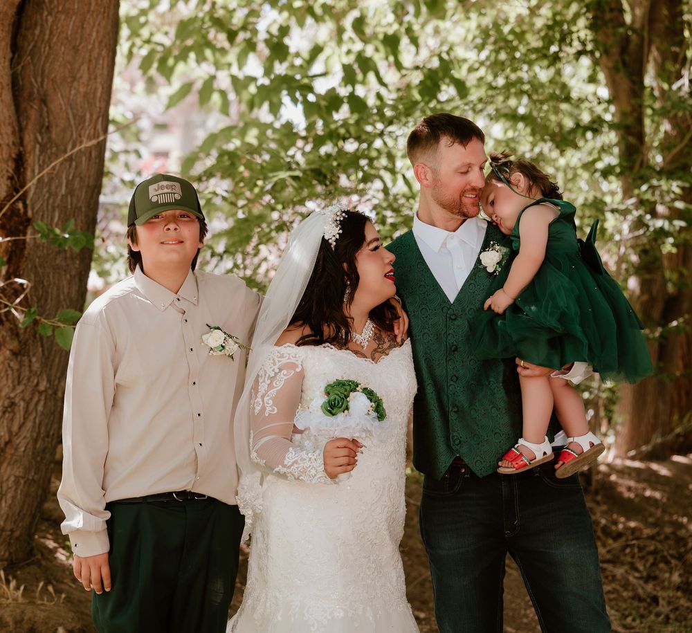 A joyful wedding family portrait with bride in lace gown, groom in green vest, teen son, and young daughter in forest.