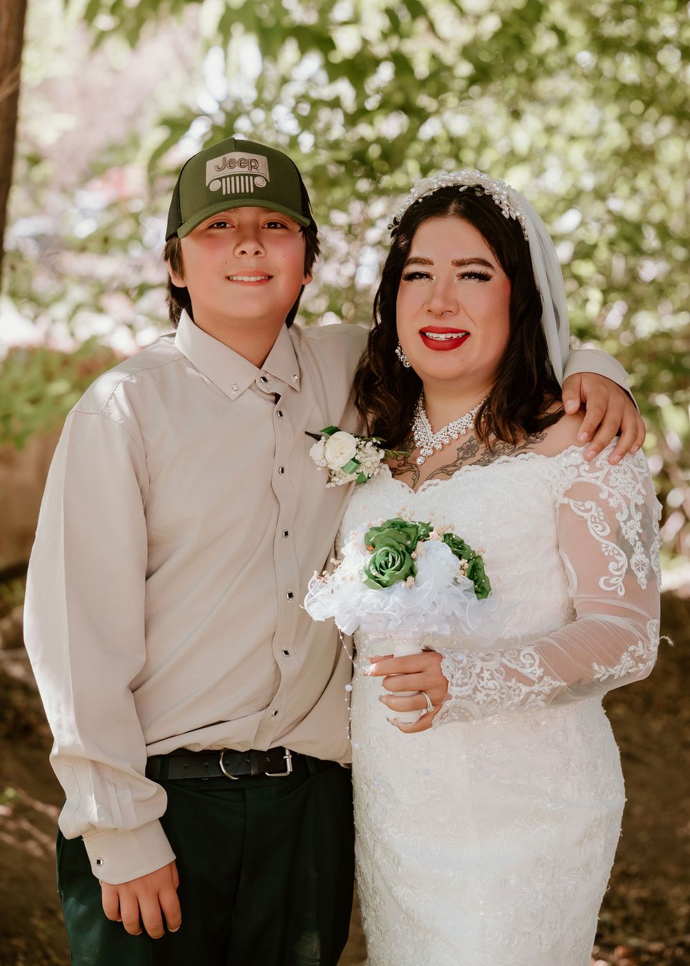 Bride in white lace gown holding green bouquet poses with young man in green cap at outdoor wedding.