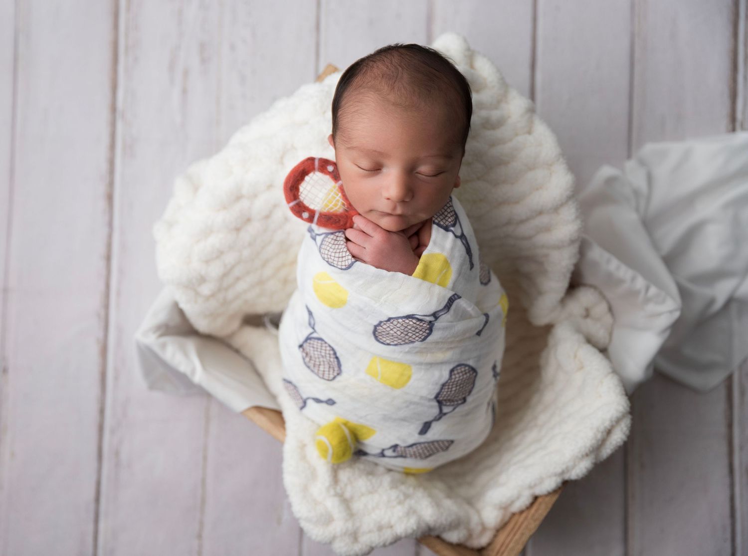 Newborn baby swaddled in a white muslin wrap with fruit print, sleeping peacefully in a wooden bowl prop.