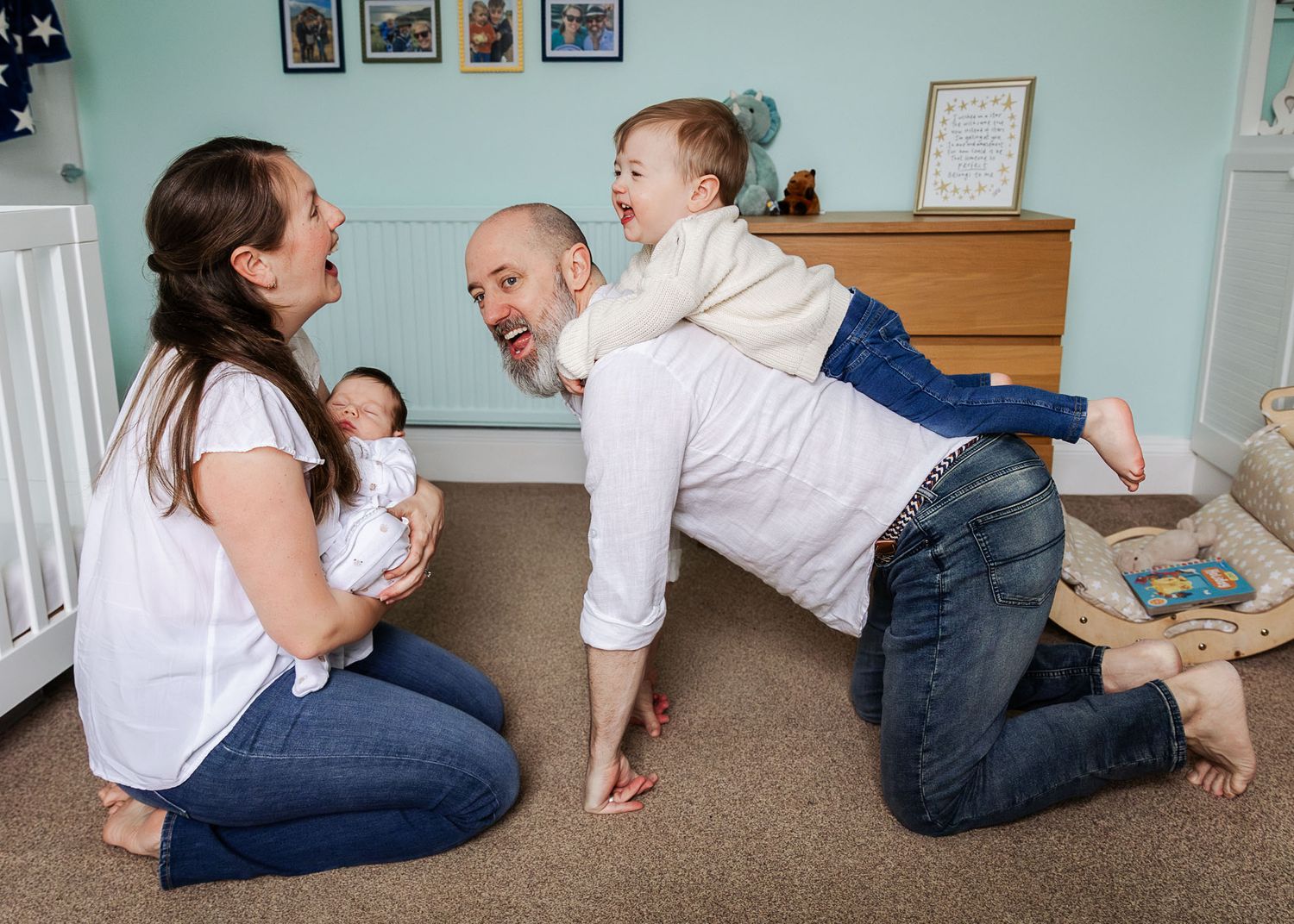During a relaxed Bristol in home photoshoot, a family laugh as mum holds her newborn son and dad gives the toddler a piggyback ride.