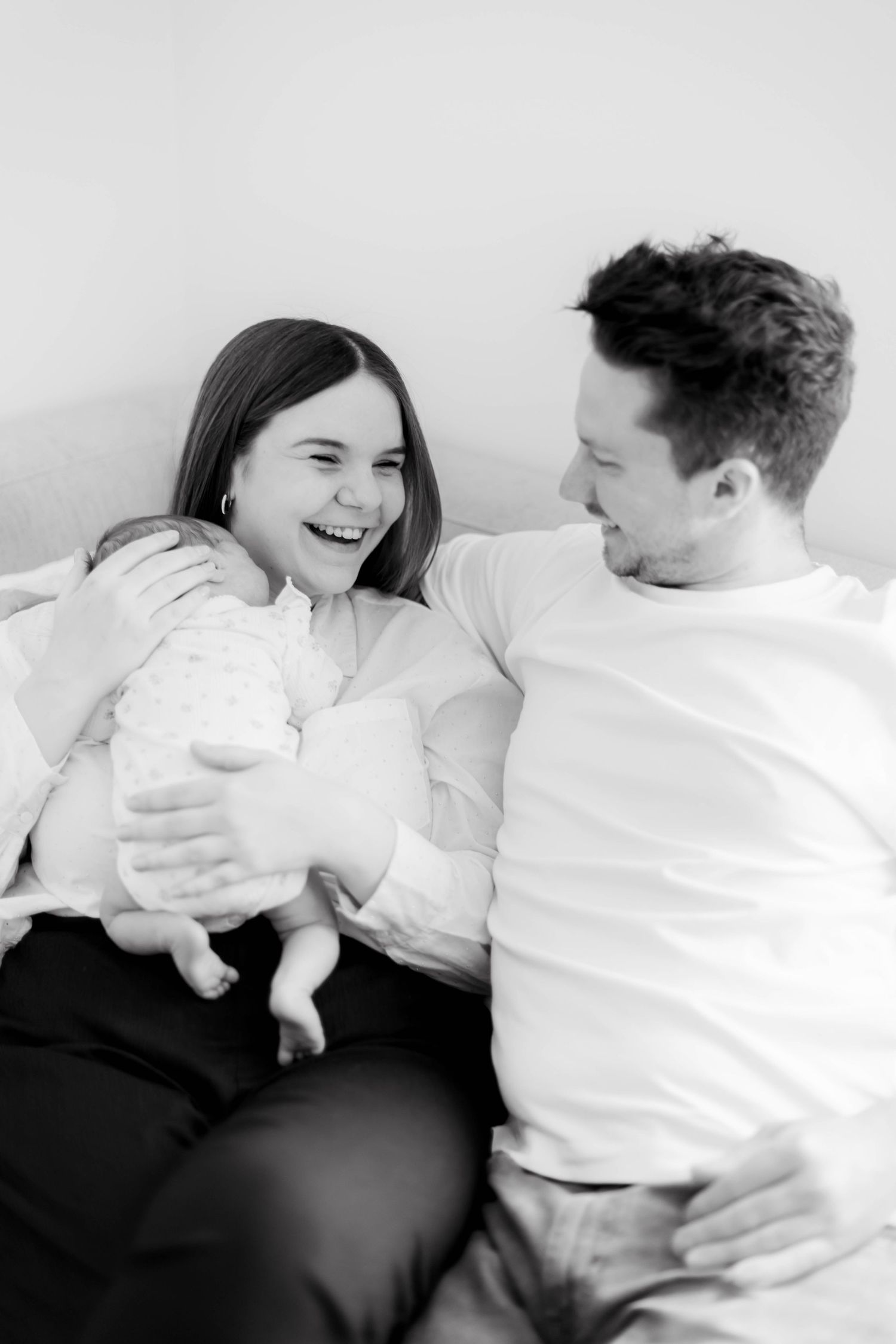Black and white photo of happy parents smiling at each other while mother holds their newborn baby on a bed.