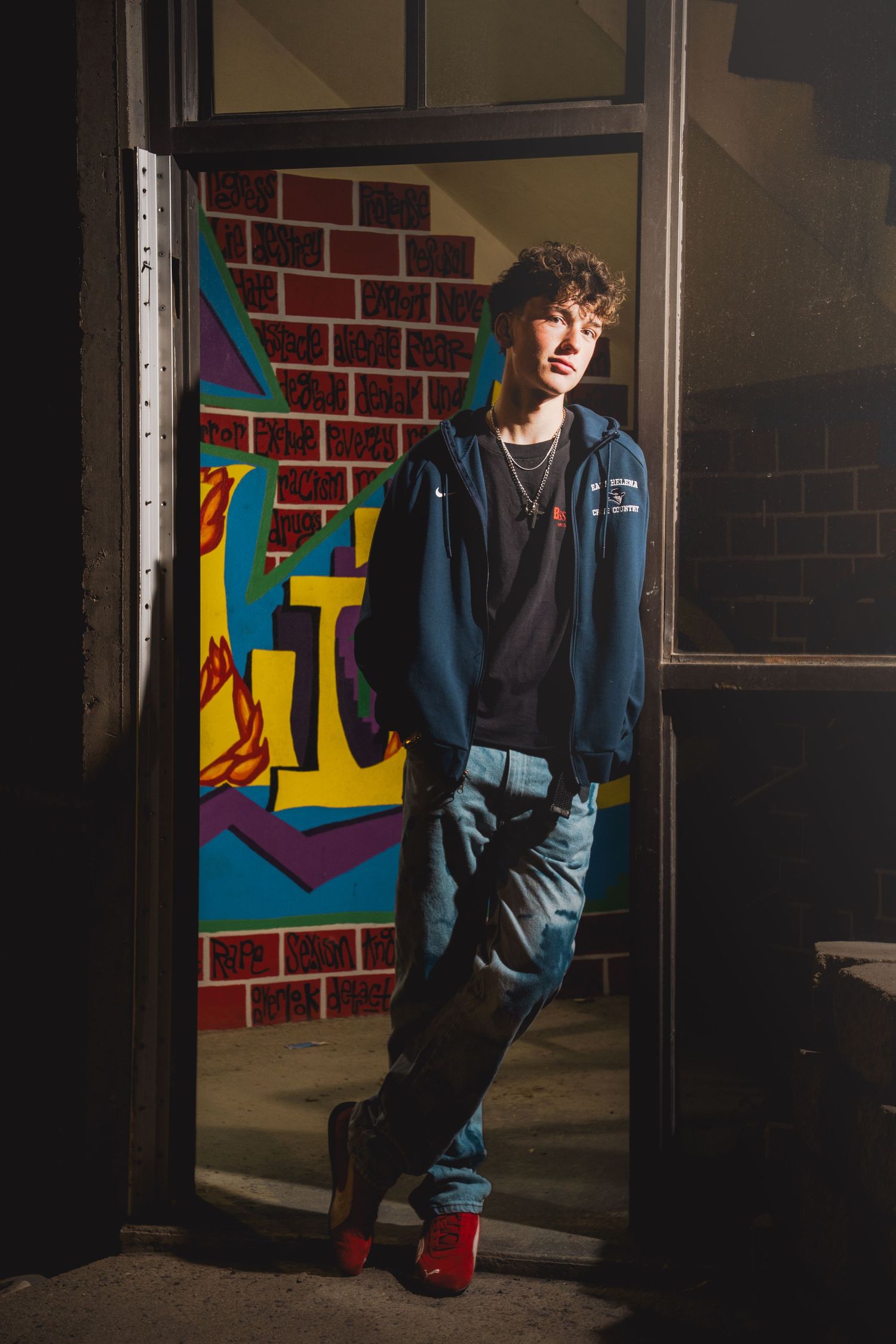 Young man in blue jacket and jeans leans against brick wall with colorful mural in doorway.