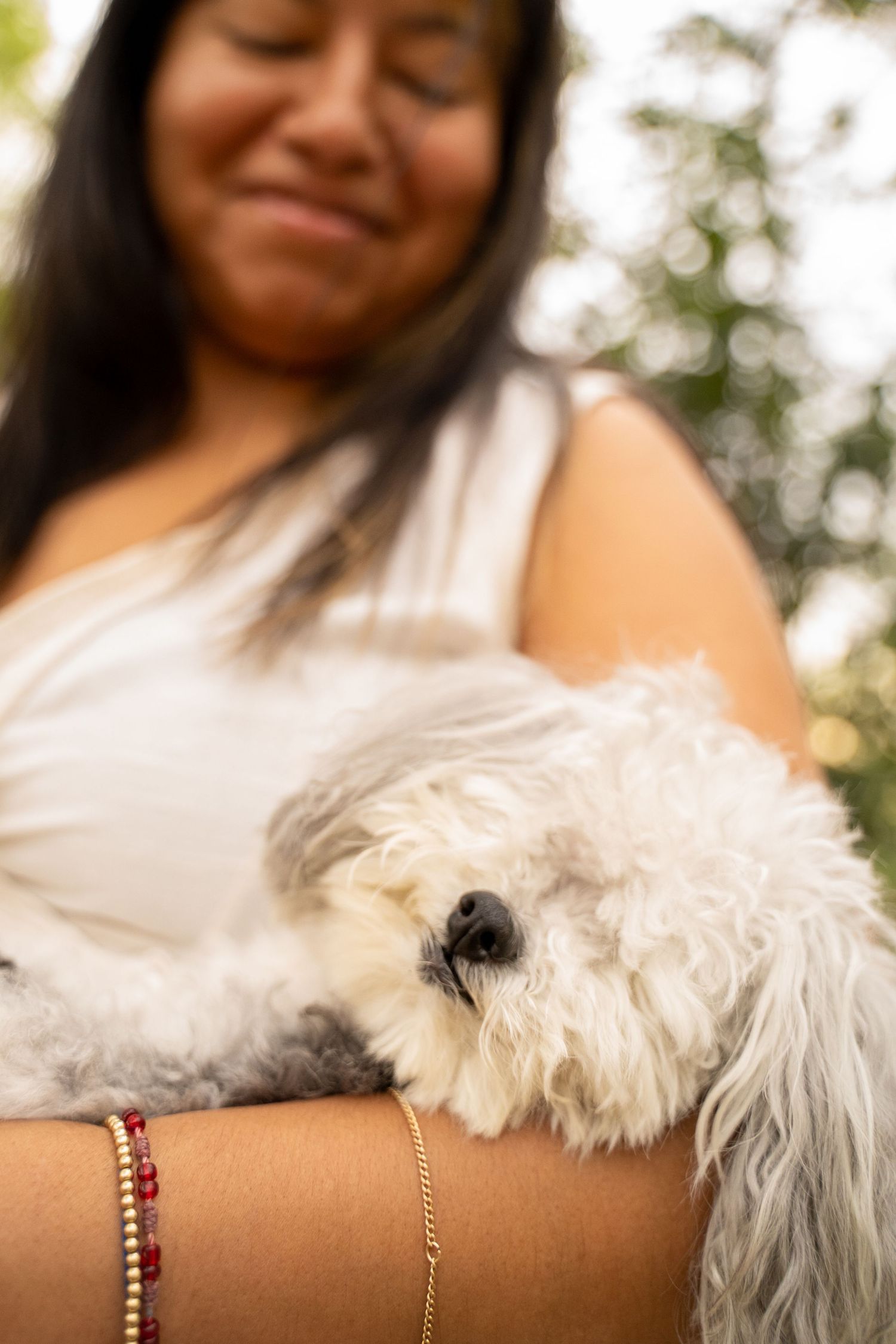 A smiling woman in a white top gently cradles a fluffy white Bichon Frise dog in her arms outdoors.