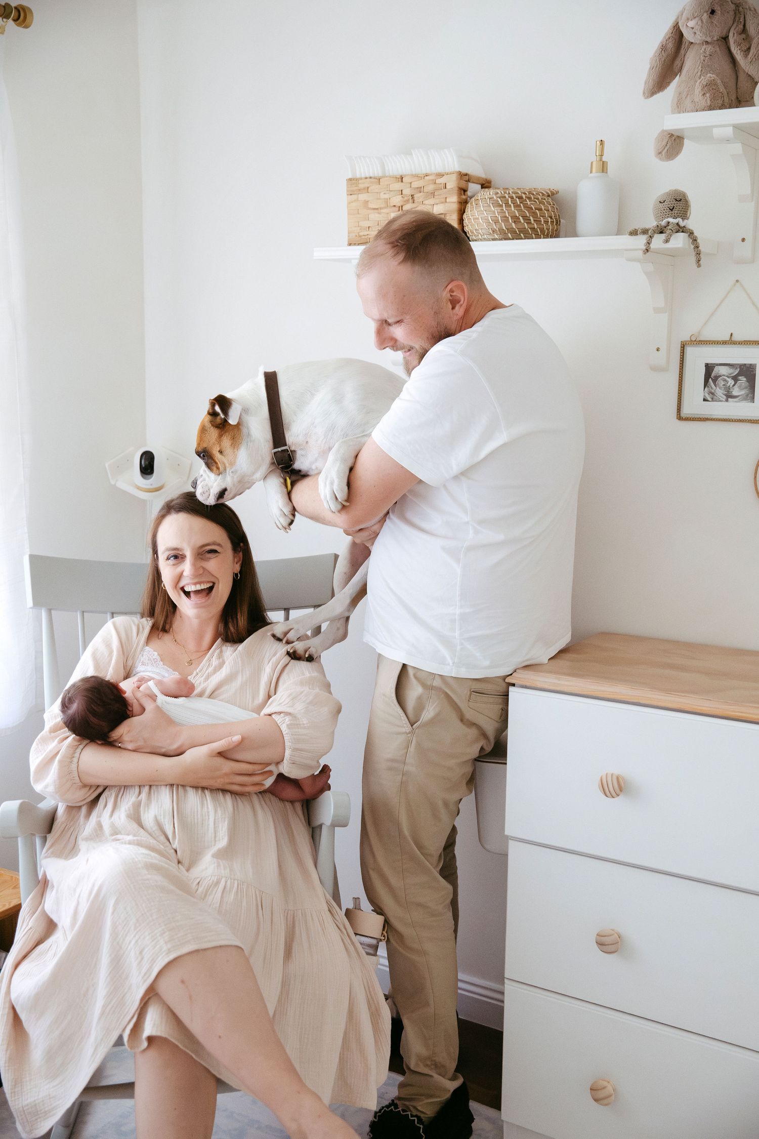 A joyful couple in a nursery, mother holding newborn while father lifts a small dog, surrounded by soft decor.
