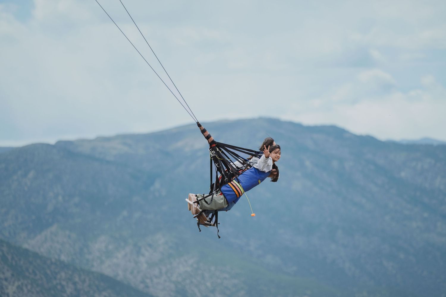 Person zip-lining over mountain landscape, suspended in harness against cloudy sky with scenic peaks in background.