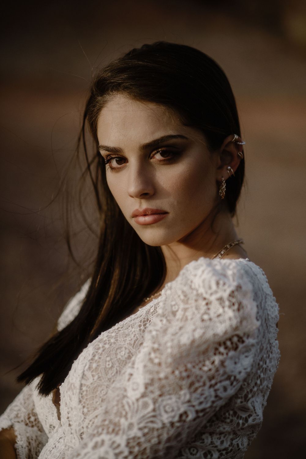 Young woman in white lace dress with ear cuff and natural makeup poses elegantly in warm golden light.