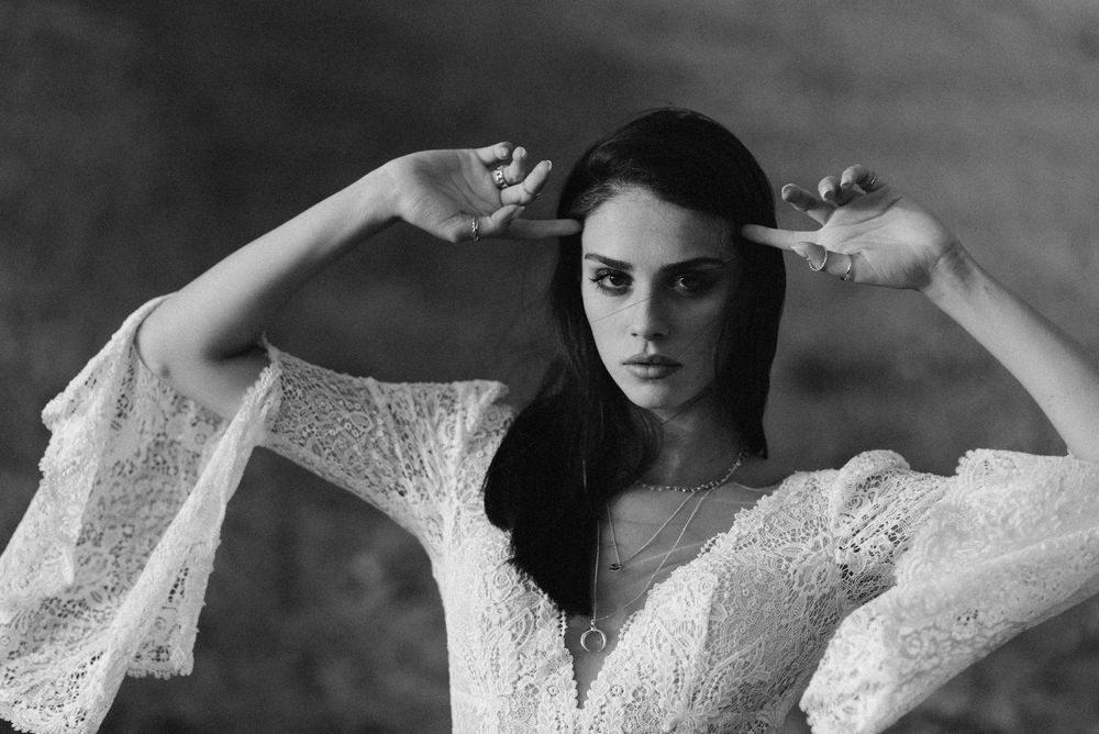Black and white portrait of a young woman in lace dress posing with hands near head outdoors.