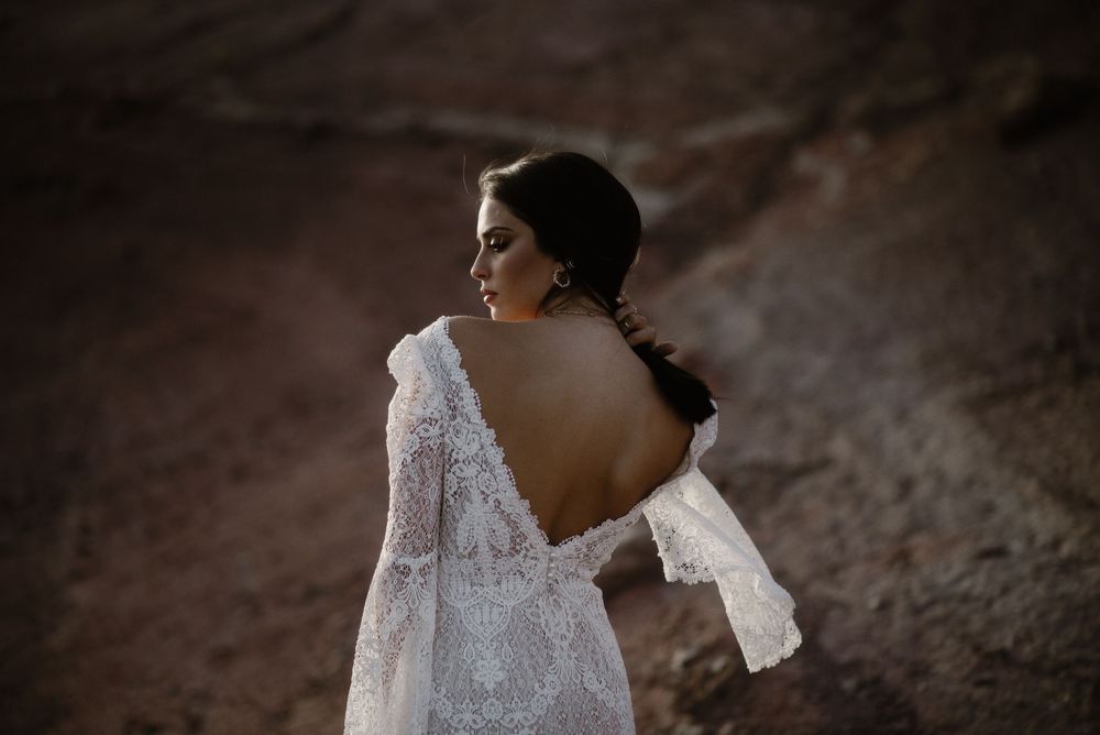 Elegant bride in backless lace wedding gown with bell sleeves stands in rocky desert setting.