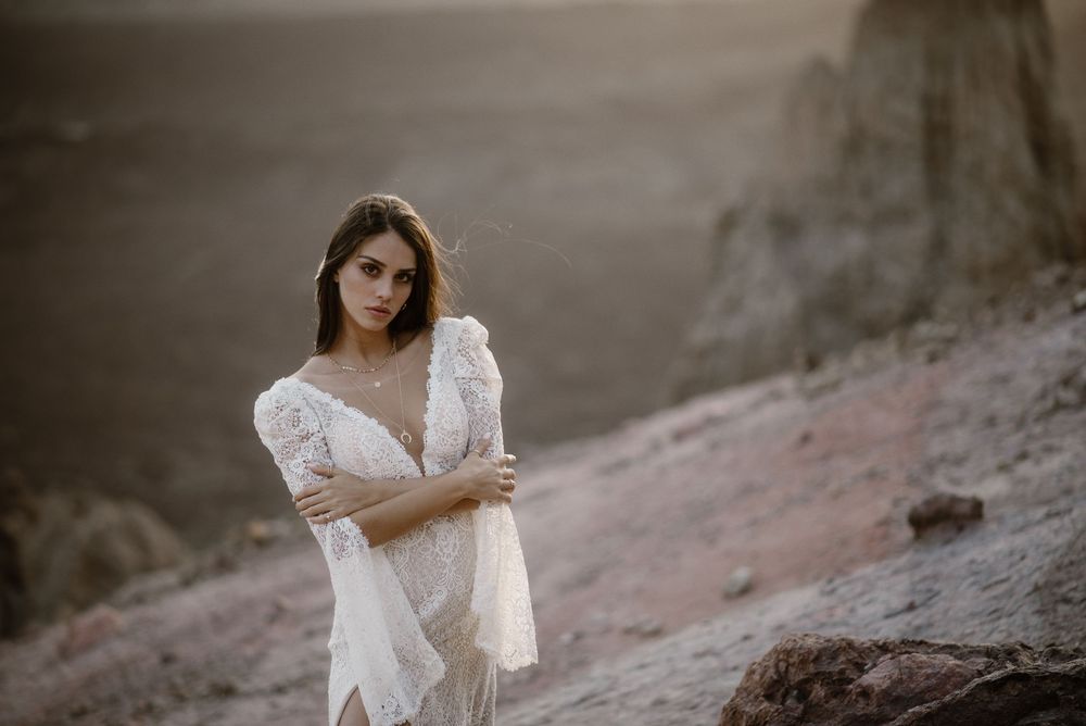 Elegant woman in white lace gown standing on rocky desert terrain at sunset.