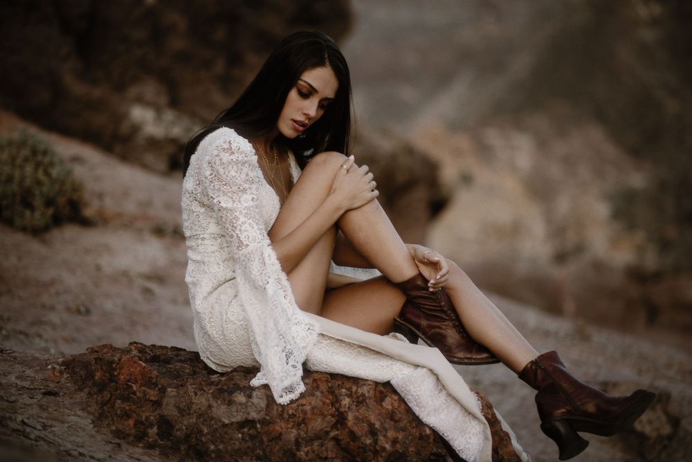 Young woman in white lace dress with knee-high socks and brown boots sitting on rocks in nature.