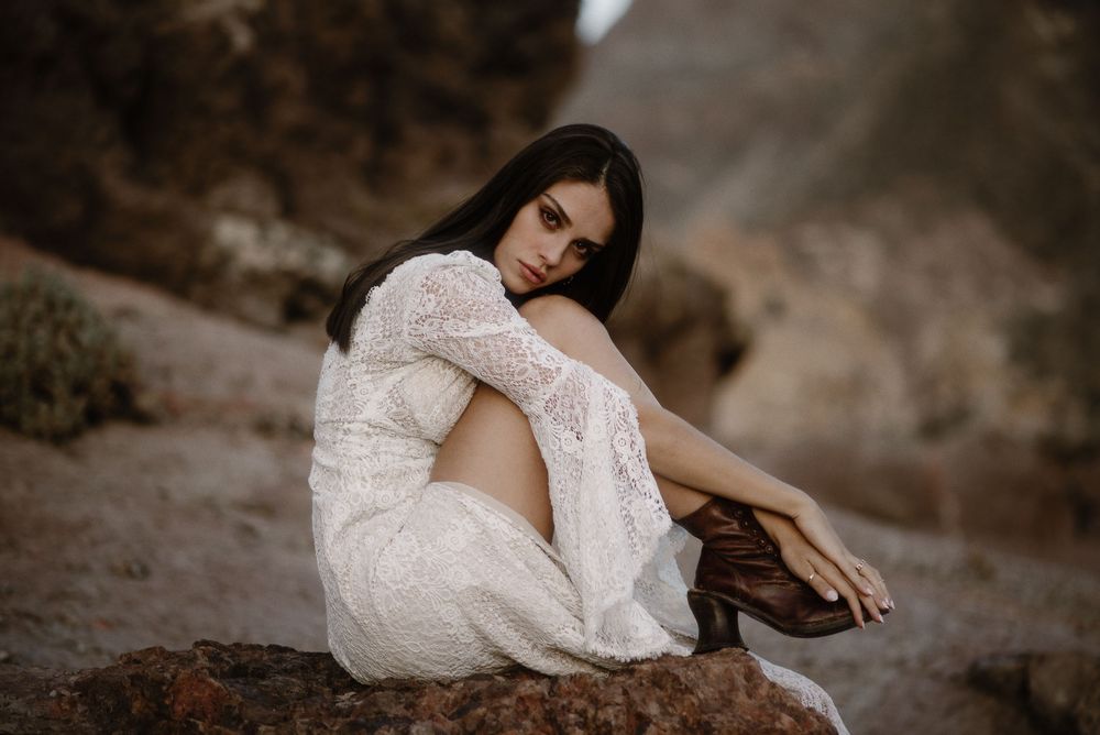 Brunette woman in white lace dress sits pensively on rocks in a rugged natural landscape.