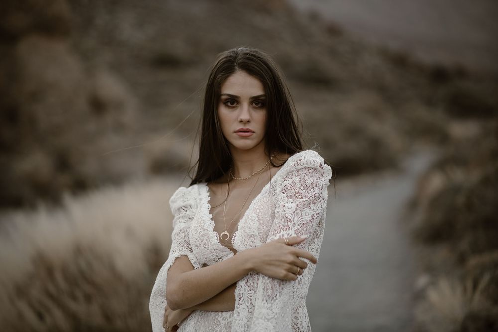 Young woman in white lace dress posing on a mountain path with arms crossed.