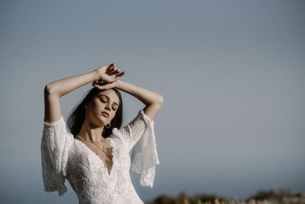 Bride in lace bell-sleeve wedding dress raises arms gracefully against soft blue sky outdoors.